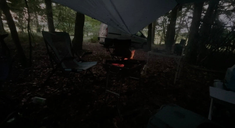 Aftenlejr i skoven med bål under tarpen og campingstole i mørket